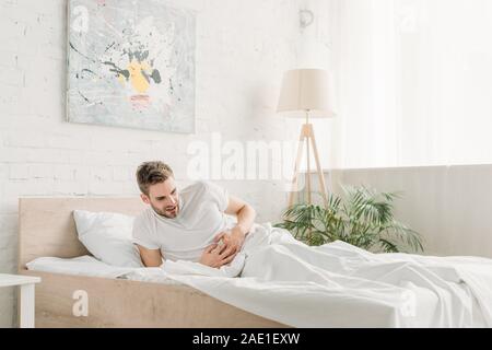 Giovane uomo toccando stomaco mentre soffrono di dolore addominale in camera da letto Foto Stock