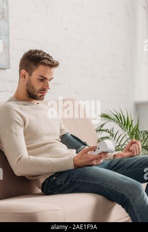 Giovane uomo seduto sul divano di casa e mediante tonometro Foto Stock