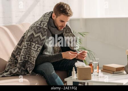 Uomo malato, avvolto in una coperta, seduto sul divano e guardando il termometro Foto Stock