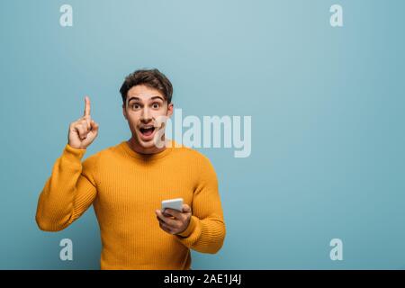 Sorpreso l'uomo utilizza lo smartphone e rivolta verso l'alto, isolato su blu Foto Stock