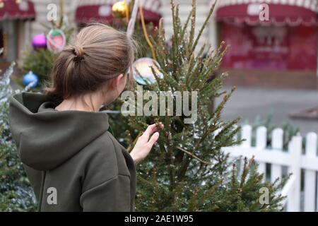Ragazza toccando albero di Natale su una strada coperta di neve. Concetto di nuovo anno celebrazione, vacanze invernali in città, compratore presso il mercato di Natale Foto Stock