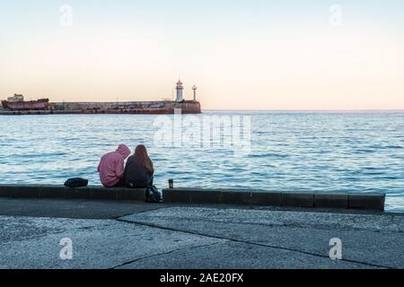 Un giovane in amore seduto sulla spiaggia, guardando il vecchio faro in distanza. Vista della costa di sera al tramonto. Foto Stock