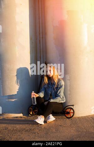 Bella donna bionda su scooter vicino a colonne di grigio al tramonto può contenere una bottiglia di acqua a portata di mano Foto Stock