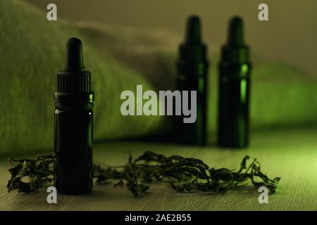 Composizione orizzontale di tre 10 ml nero bottiglie di vetro circondato da erbe essiccate su bianco tavolo in legno Retroilluminato con luce verde Foto Stock