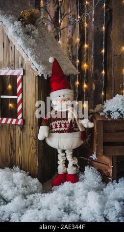 Un grande pupazzo di neve in una Red Hat e sciarpa bianco, in un elegante luce rossa, sorge nei pressi di una casa in legno su un inverno sfondo di Natale. Decorazioni in una foto st Foto Stock