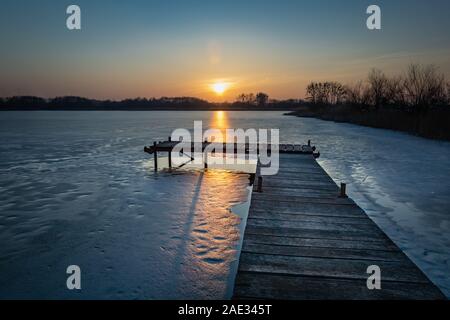 Ponte di legno fatto di schede, lago ghiacciato e al tramonto, i trend con orizzonte di riferimento e la luce sul riquadro di ghiaccio Foto Stock