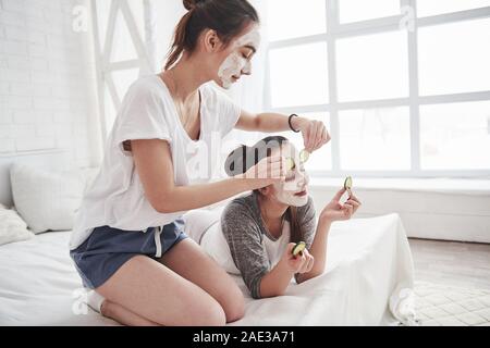 Giornata di bellezza. Concezione della cura della pelle utilizzando la maschera bianca e i cetrioli sul viso. Due sorelle femmina hanno weekend in camera da letto Foto Stock