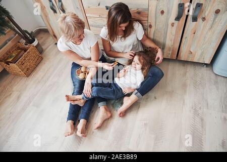 Kid giacente sulla capogruppo per le gambe. Madre, la nonna e la figlia avente un buon tempo in cucina Foto Stock