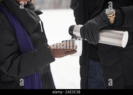 Vista ravvicinata dell'uomo versando il tè caldo da thermos per la tazza nella donna le mani in inverno al di fuori Foto Stock