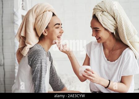 Asciugamani sulle teste. Concezione della cura della pelle utilizzando la maschera bianca sul viso. Due sorelle femmina hanno weekend in camera da letto Foto Stock