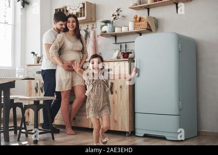 La famiglia felice. Femmina giocoso bambino divertirsi eseguendo in cucina al giorno di fronte a lei il padre e la madre Foto Stock