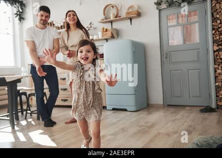 Con la lingua di fuori. Femmina giocoso bambino divertirsi eseguendo in cucina al giorno di fronte a lei il padre e la madre Foto Stock