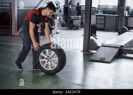 In rosso e colorati di nero uniforme. Mechanic tenendo un pneumatico in corrispondenza del garage di riparazione. Sostituzione delle gomme invernali ed estive Foto Stock