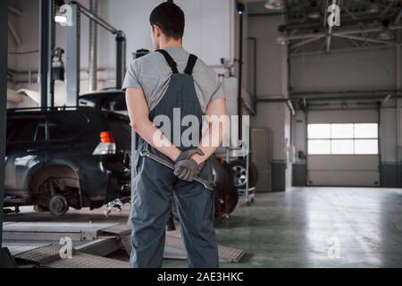 Wathing all'auto. Vista posteriore del lavoratore in uniforme che si erge in officina con la chiave in mano Foto Stock