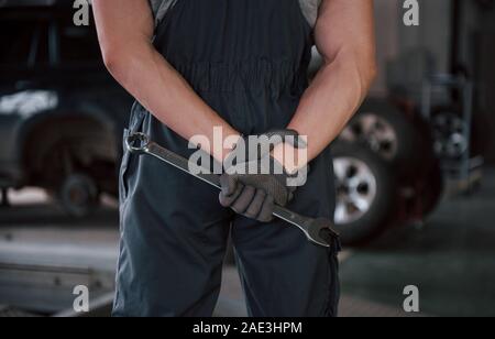 Mechanic guarda avanti. Vista posteriore del lavoratore in uniforme che si erge in officina con la chiave in mano Foto Stock