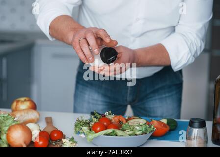 Mettere un po' di sale. Uomo in camicia bianca preparare del cibo su la cucina a base di verdure Foto Stock