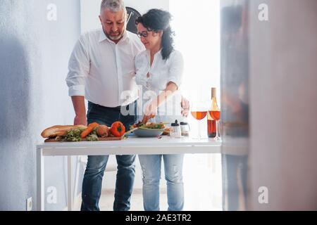 Essi ancora giovane. L'uomo e sua moglie in una camicia bianca preparare del cibo su la cucina a base di verdure Foto Stock