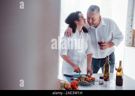 Unshaved marito dando un po' d'amore. L'uomo e sua moglie in una camicia bianca preparare del cibo su la cucina a base di verdure Foto Stock