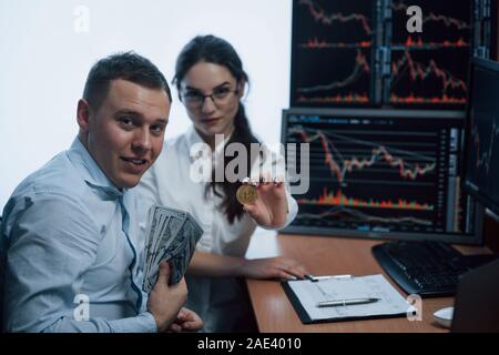 Mostra off guadagnato le cose. Bella donna e uomo allegro holding bitcoin e denaro nelle mani mentre è seduto in un ufficio moderno con molti monitor con Foto Stock