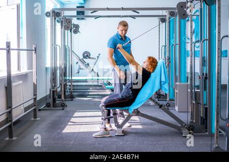 Personal trainer assiste la donna con disabilità nel suo allenamento. Sport centro riabilitativo con fisioterapisti e pazienti che lavorano insieme verso hea Foto Stock