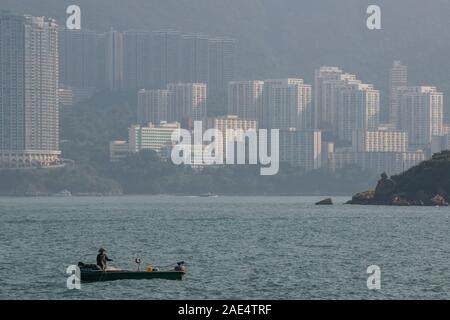 Un pescatore commerciale su una piccola barca da pesca con lo sfondo di edifici dell'Isola di Hong Kong con il porto di Aberdeen Foto Stock Un pescatore commerciale su una piccola barca da pesca con lo sfondo di edifici dell'Isola di Hong Kong con il porto di Aberdeen Foto Stock