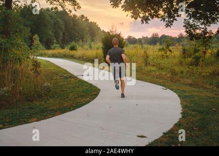 Vista posteriore di un maschio di atletica che corre lungo un percorso in un parco al tramonto Foto Stock