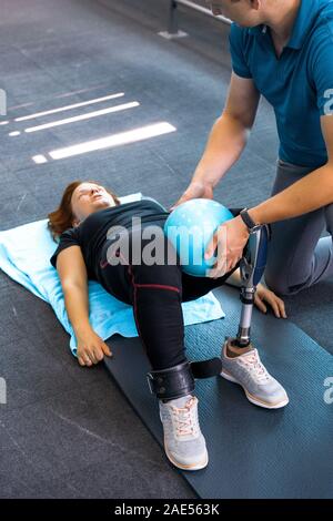 Personal trainer assiste la donna con disabilità nel suo allenamento. Sport centro riabilitativo con fisioterapisti e pazienti che lavorano insieme verso hea Foto Stock