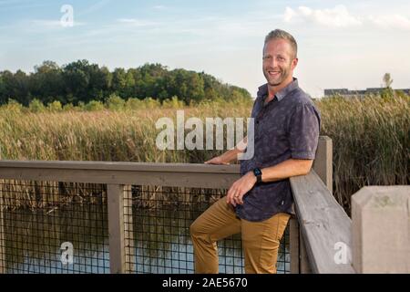 Un bel uomo sorridente si appoggia contro una ringhiera in un parco di zone umide Foto Stock