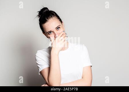 Affascinante giovane donna sinceramente ride, chiude la bocca con la mano e guarda la telecamera allegramente. Foto Stock