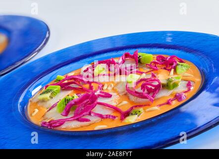 Vista frontale del carpaccio di merluzzo con crema di pomodoro, cavolo rosso, kiwi e Tostate i pinoli in una piastra di colore blu su sfondo bianco. Foto Stock