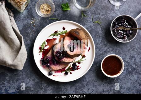 Gustose grigliate di carne di manzo con salsa di mirtilli sulla piastra bianca su pietra blu sullo sfondo. Mezzo raro arrosto di manzo con salsa di frutti di bosco. Vista superiore, laici piatta Foto Stock