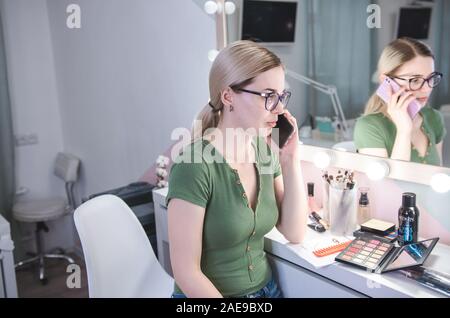 Femmina bionda make up artist parla al telefono con il cliente. Parrucchiere professionista nella sua bellezza visage studio Foto Stock