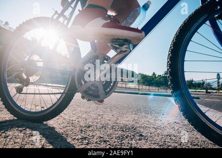 Montare maschio ciclista biker in sella alla sua moto ciclo su una strada asfaltata al tramonto mentre il sole tramonta attraverso la sua ruota. Foto Stock