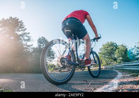 Montare maschio ciclista biker in sella alla sua moto ciclo su una strada asfaltata al tramonto mentre il sole tramonta attraverso la sua ruota. Foto Stock