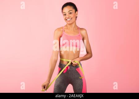 Montare ragazza afro cintura di misurazione con nastro in piedi, sfondo rosa Foto Stock