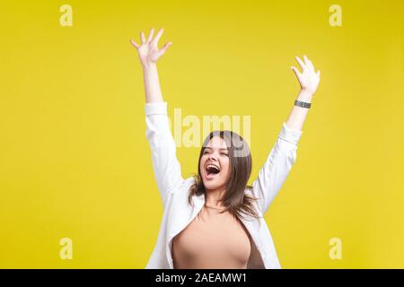 Ritratto di estremamente felice bella bruna donna in giacca e jeans alzando le mani e urlando con entusiasmo, celebrando la vittoria di successo ind. Foto Stock