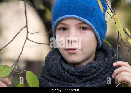 Pretty Boy bambino indossare caldo abbigliamento invernale azienda ramo di albero con foglie verdi in tempo freddo all'esterno. Foto Stock