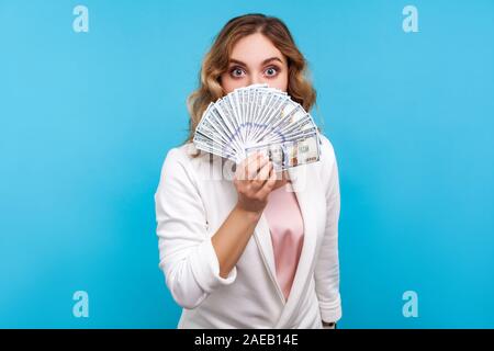 Ritratto di giovane bella donna con capelli ondulati e sorpreso grandi occhi indossando giacca bianca che copre metà faccia con banconote di dollari, grande profitto. in Foto Stock