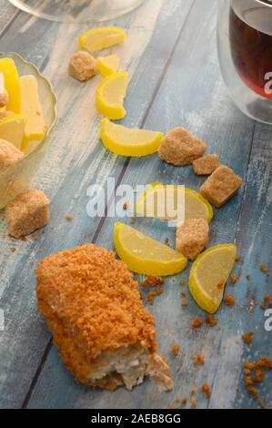 Uno rotto torta, un bicchiere di tè nero, pezzi di zucchero di canna e marmellata fette di limone su un sfondo di legno Foto Stock