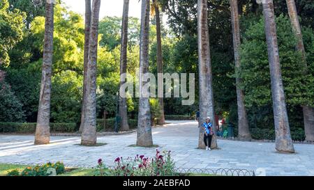 Donna asiatica con fedora bianco e cravatta blu-morto t-shirt in piedi da palme in Giardini Nazionali a Atene, Grecia Foto Stock