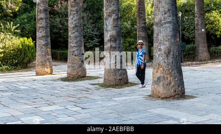 Donna asiatica con fedora bianco e cravatta blu-morto t-shirt in piedi da palme in Giardini Nazionali a Atene, Grecia Foto Stock