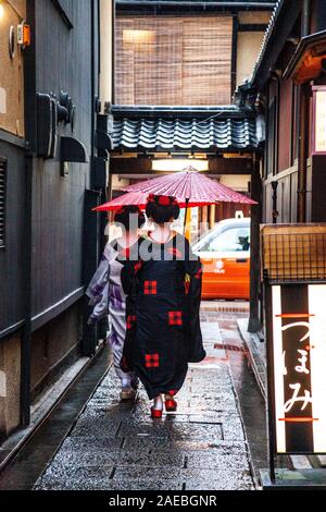 Due geisha / maiko camminando per uno stretto vicolo in possesso di una carta di riso parasol nella piovosa quartiere Gion, Kyoto, Giappone Foto Stock