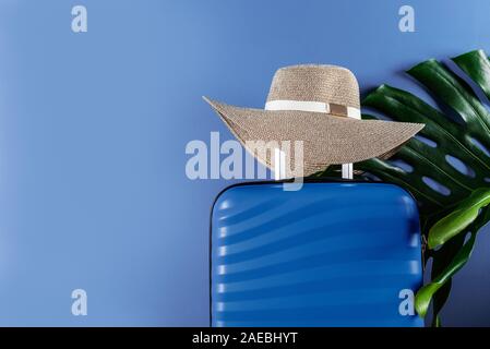 Un elegante e luminoso blu dimensioni cabina valigia con cappello di paglia e monstera foglia contro sfondo blu scuro. Facile viaggiare con poco bagaglio concetto. Copia dello spazio. Foto Stock