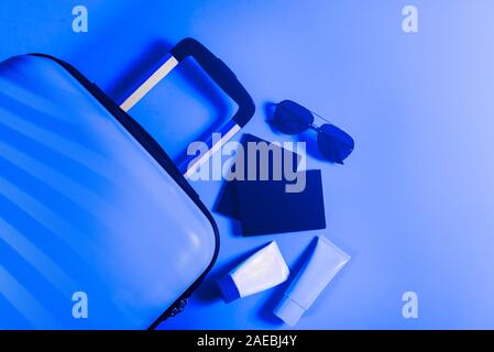 Un elegante e luminoso dimensioni cabina valigia con cappello di paglia, protezione solare e passaporti laici piana. Facile viaggiare con poco bagaglio concetto. Copia dello spazio. Tonica nell'immagine blu Foto Stock