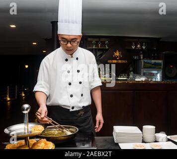 Uomo asiatico che indossa cappello e grembiule da chef sorride come lui  guarda le ciambelle sul tavolo mentre scrivi un notebook mentre si è seduti  in cucina Foto stock - Alamy