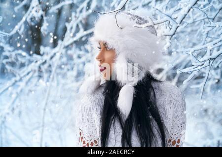 Bel modello in elegante cappello caldo in posa nella foresta vicino a rami di albero con neve, guardando lontano. Bellissima ragazza con lunghi capelli neri in bianco moderno abbigliamento. Foto Stock