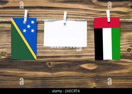 Appendere bandiere delle Isole Salomone e Emirati Arabi Uniti attaccato alla corda con vestiti perni con copia spazio bianco sulla nota carta su sfondo di legno. Foto Stock