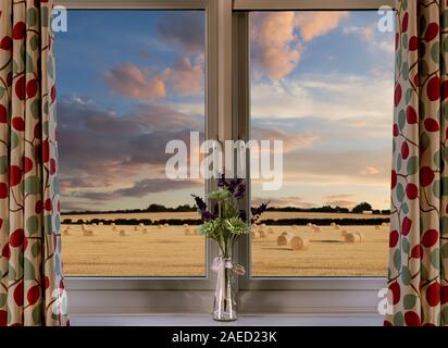 Guardando fuori da una bellissima finestra su un campo rurale di grano e balle di fieno durante il tramonto Foto Stock