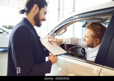 Una giovane donna acquista un'auto firma un contratto in una sala di esposizione. Un uomo firma un contratto di autonoleggio. Foto Stock