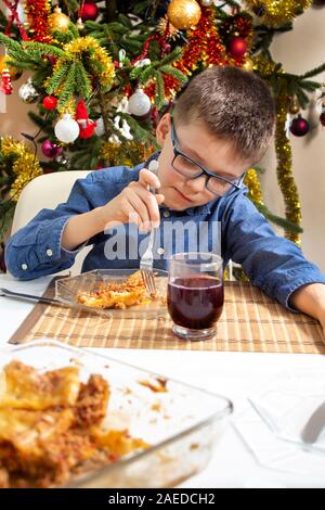 Il ragazzo con gli occhiali si siede al tavolo e il cibo di poke sulla sua forcella sulla piastra. Un bellissimo albero di Natale decorato in background. Foto Stock